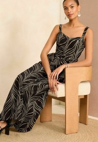 Fekete maxi ruha fehér hullámos mintákkal, négyzet alakú dekoltázzsal és vastag pántokkal. A modell egy világos fa székben ül, amelynek puha ülése van.