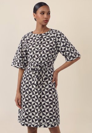 Frau trägt ein knieumspielendes Kleid mit schwarz-weißem geometrischem Muster, halblangen Ärmeln und einem gebundenen Taillengürtel, steht mit einer Hand in der Hüfte.