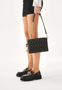 Black handbag with quilted texture, gold chain accents, and detachable strap. Worn with black patent loafers featuring chain detail.