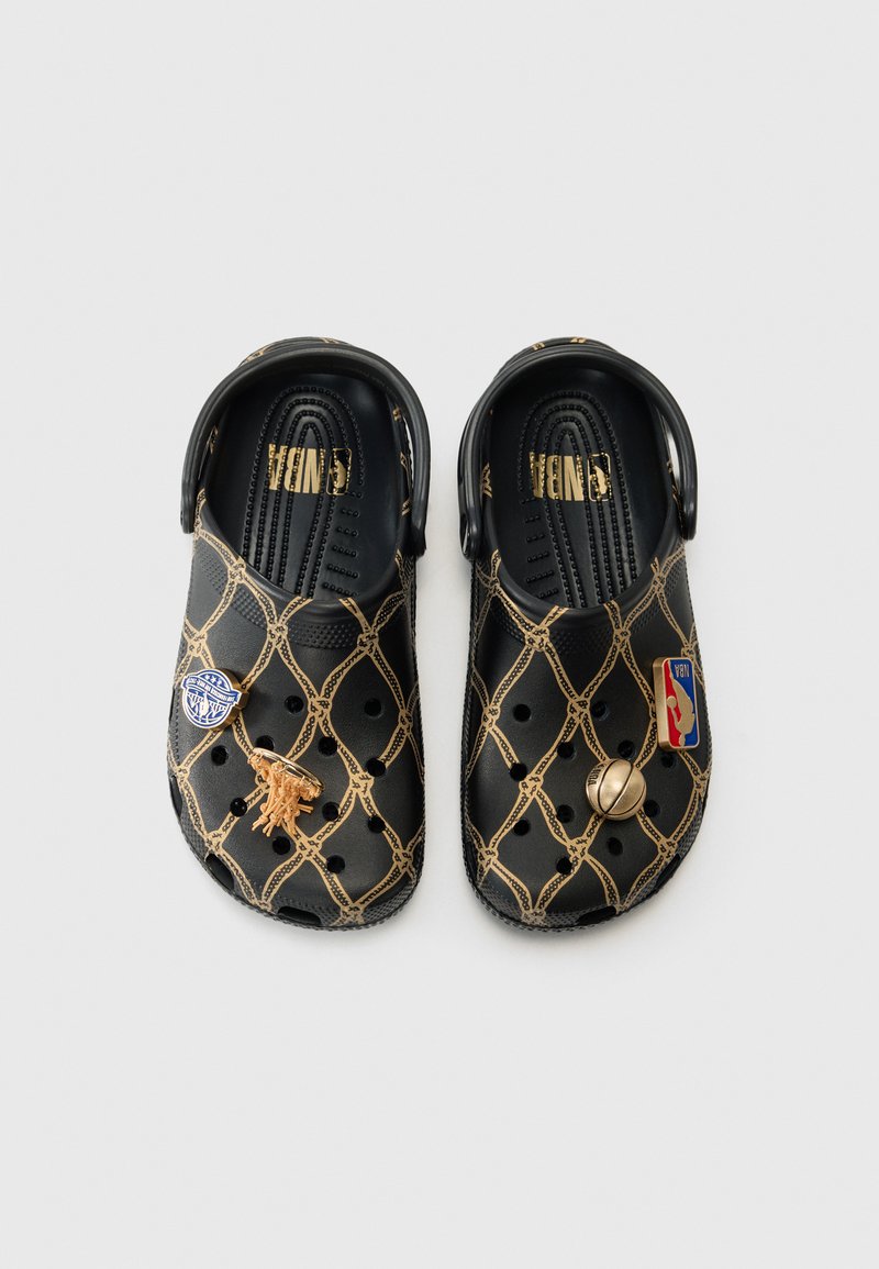 Schwarze Crocs mit einem goldenen Kettenmuster, versehen mit drei dekorativen Pins: einem Löwen, einem Basketball-Logo und einem runden Akzent an der Vorderseite.