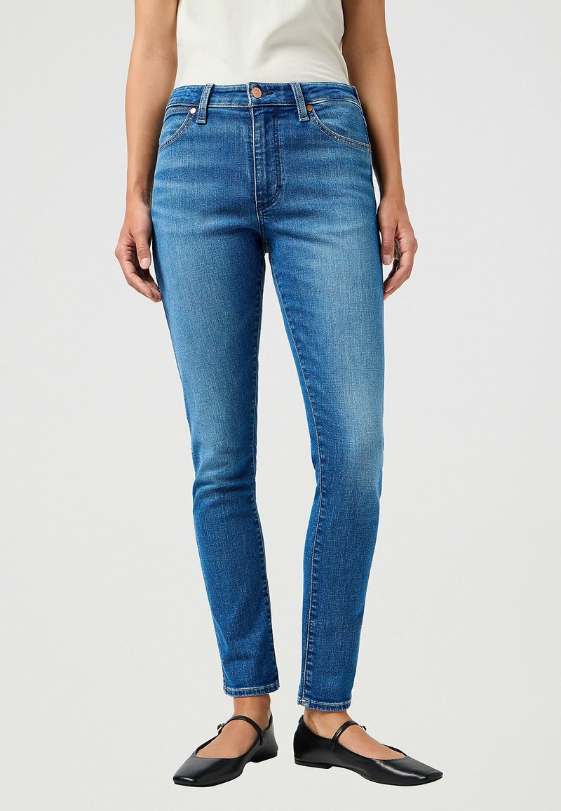 Femme portant un jean slim bleu, un haut blanc et des ballerines Mary Jane noires, debout devant un fond clair uni.