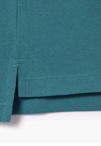 Teal strikket stof, der viser en nærbillede af en garments kant med sideslids og dobbeltsyet detalje.