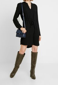 Schwarzes langärmliges Kleid mit V-Ausschnitt und Knotenbund, kombiniert mit kniehohen olivgrünen Texturstiefeln und einer kleinen marineblauen Umhängetasche.