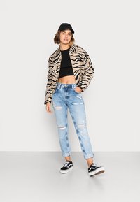 Jaqueta bomber em padrão de zebra, top curto preto, jeans claros rasgados,sapatos pretos com atacadores brancos e boné preto. Fundo simples.