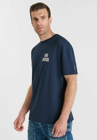 Man in een marineblauw T-shirt met de tekst "BIG BOSS" op de borst en blauwe denim jeans, staand tegen een effen witte achtergrond.