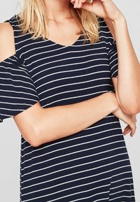 Kvinna som bär en marinblå och vit randig klänning med kallaxlar och korta ärmar, med armarna i kors och handen n ära ansiktet.