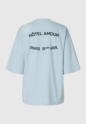 T-shirt ample bleu clair avec le texte noir « HÔTEL AMOUR » et « PARIS 9ÈME ARR. » imprimé au dos.