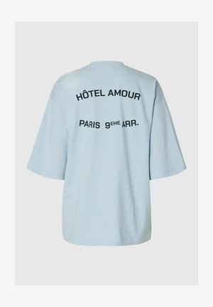Heleroheline oversize-t-särk, mille seljal on musta värvi tekst "HÔTEL AMOUR" ja "PARIS 9ÈME ARR.".