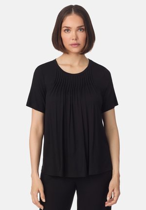 Femme aux cheveux bruns foncés et raides, portant un haut noir à manches courtes avec un détail plissé à l'avant, sur un fond uni.