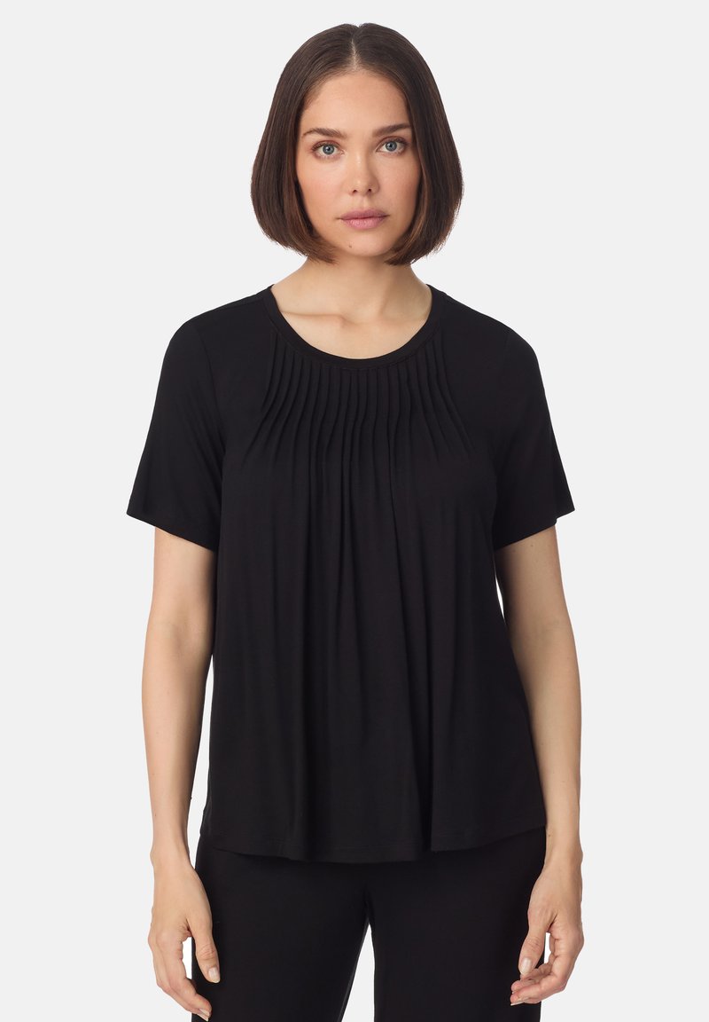 Frau mit glattem dunkelbraunem Haar, die ein schwarzes Kurzarmoberteil mit plissiertem Vorderteil vor einem einfarbigen Hintergrund trägt.