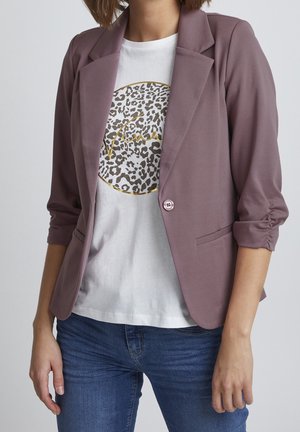Femme portant un blazer mauve aux manches froncées par-dessus un t-shirt blanc à imprimé léopard et un jean bleu, debout devant un fond uni.