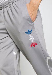 Osoba oblečená v světle šedých teplákových kalhotách s modrými, bílými a červenými trefoil logy Adidas na nohavici, ruka v boční kapse.