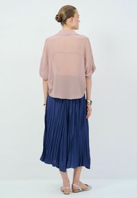 Blusa leggera rosa chiaro con maniche corte a dolman, abbinata a una gonna fluida e plissettata blu navy. Il modello è in piedi di spalle alla fotocamera.