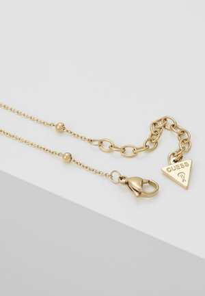Gouden ketting met kreeftsluiting, verstelbare schakels en een driehoekige bedel gegraveerd met "GUESS" en een vraagteken.