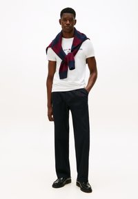 Valge T-särk, tumesinised püksid, mustad kingad; tumesinine ja burgundia villa sall õlgadel; lihtne taust.