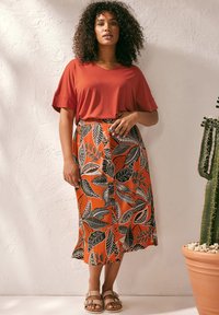 Top rojo de manga corta con escote en V combinado con una falda naranja que presenta grandes patrones de hojas negras. Sandalias simples completan el conjunto.
