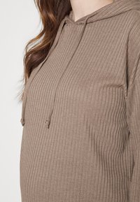 Primo piano di una donna incinta che indossa una felpa a coste di colore marrone chiaro con coulisse, che mostra il suo pancione e i capelli sciolti.