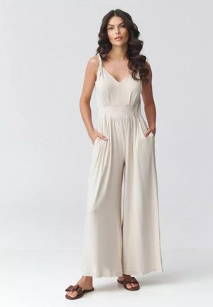 Kobieta o długich ciemnych włosach, ubrana w bezrękawnikowy beżowy kombinezon i brązowe sandały, stojąca z rękami w kieszeniach na jednolitym tle.