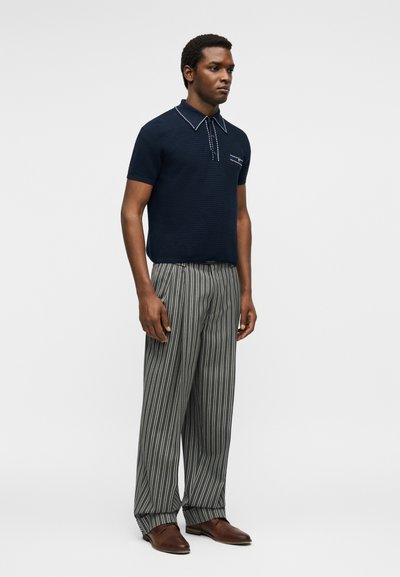 Homme debout portant un polo en maille bleu marine à manches courtes, un pantalon large rayé gris et blanc, et des chaussures en cuir marron sur fond blanc.