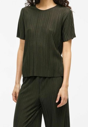 Femme portant un haut plissé à manches courtes vert foncé et un pantalon large plissé assorti, debout devant un fond blanc uni.