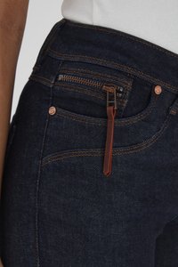 Közeli kép sötétkék farmernadrágról narancssárga varrással, vörösréz gombbal és egy kis cipzáros zsebbel, barna bőr húzófüllel.