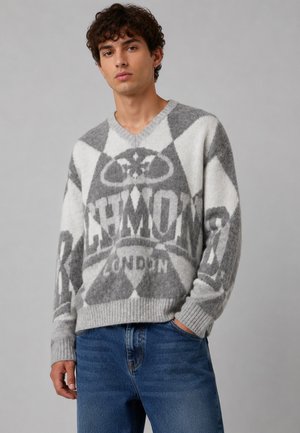 Giovane uomo con capelli ricci che indossa un maglione lavorato a maglia grigio e bianco con il testo "RICHMOND LONDON" e jeans blu su sfondo grigio.