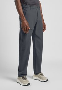 Graue Outdoor-Hosen aus leichtem Stoff mit geradem Schnitt, ausgestattet mit einer Reißverschlusstasche und Gürtelschlaufen, kombiniert mit beigen Sportschuhen.