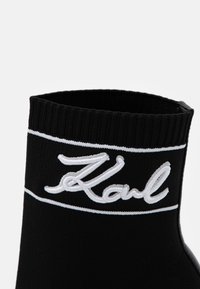 Calcetín negro de punto con un puño acanalado. Presenta un logo bordado en blanco que dice "Karl" y rayas horizontales blancas cerca de la parte superior.