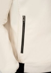 Veste en cuir blanc avec une finition texturée, détail de fermeture éclair noir sur la poche latérale et ceinture côtelée noire.