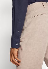 Camisa de manga larga color marino con detalles de puños abotonados. Pantalones color beige claro con una tela texturizada y bolsillo trasero. Primer plano, mostrando el ajuste y los colores.