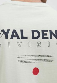 Witte katoenen t-shirt met grote zwarte tekst "ROYAL DENIM DIVISION" op de achterkant, met rode cirkelvormige grafische details en kleine tekst eronder.