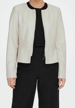 Femme portant une veste zippée beige clair sur un haut noir et un pantalon noir, debout les mains détendues le long du corps.
