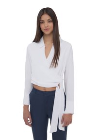 Blusa bianca avvolgente con colletto, maniche lunghe e cintura in vita. Indossata con pantaloni sartoriali blu navy. Tessuto liscio, design minimalista.