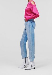 Frau in hellblauen, mit Strasssteinen verzierten Jeans, weißen, hohen Stiefeletten und einem langen, rosa Oberteil mit Schulteausschnitten, seitlich stehend.