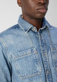 Camisa de mezclilla azul claro con cuello abotonado, dos bolsillos en el pecho y un sutil detalle de costura en el hombro izquierdo.