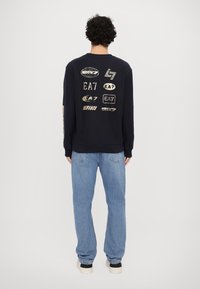 Jūras spēku krāsas džemperis ar zelta logotipa druku uz muguras, ar garām piedurknēm un apaļu kakla izgriezumu. Apvienots ar gaiši ziliem džinsiem un sporta apaviem.