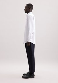 Witte blouse met lange mouwen en knoopaccenten, gecombineerd met donkere broek en zwarte schoenen. De blouse heeft een klassieke kraag en zijslits.