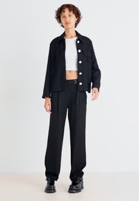 Chaqueta negra texturizada con grandes botones blancos, combinada con un top corto blanco y pantalones de pierna ancha negros, que cuentan con una cintura ajustada y botas gruesas.