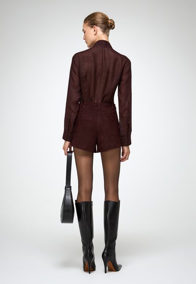 Camicia trasparente color bordeaux abbinata a pantaloni corti a vita alta di un bordeaux scuro. La modella indossa stivali neri con tacco a stiletto al ginocchio e porta una borsa nera.