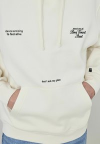 Hoodie crème en tissu doux avec trois phrases brodées en noir : "danse et chante pour te sentir vivant", "ne demande pas mon plan", et "retrouve-moi rue Bon Vivant."
