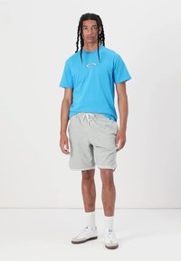 Modra kratka majica s kratkimi rokavi z logotipom, svetlo sivi športni kratki hlači z vrvico, bele nogavice in bele superge z rjavimi akcenti.