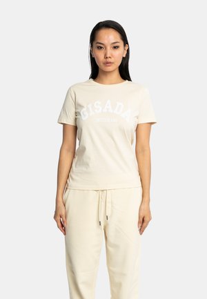 Femme portant un t-shirt beige Gisada Switzerland et un pantalon à cordon assorti, debout devant un fond uni gris clair.