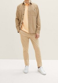 Beige kord skjorta, ljus orange pikétröja under, ljus beige byxor och vita sneakers. Två bröstfickor på skjortan.