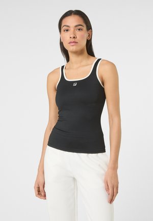 Kobieta ubrana w dopasowany czarny tank top z białym obszyciem oraz białe spodnie, stojąca na jednolitym jasnoszarym tle.