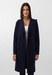 Cappotto lungo blu navy con cappuccio, realizzato in un tessuto testurizzato. Presenta due tasche frontali e un design minimalista, ideale per essere indossato a strati.