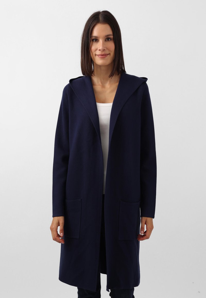 Cappotto lungo blu navy con cappuccio, realizzato in un tessuto testurizzato. Presenta due tasche frontali e un design minimalista, ideale per essere indossato a strati.