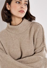 Beige gestrickter Rollkragenpullover mit geripptem Kragen und Ärmeln. Weicher Stoff und entspannter Schnitt mit detaillierten Nahtakzenten an den Schultern.