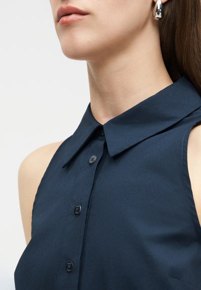 Νέα γυναίκα που φοράει ασημένιο σκουλαρίκι σε σχήμα δάκρυ και αμάνικο μπλε σκούρο πουκάμισο με γιακά και κουμπιά, μπροστά από απλό φόντο.