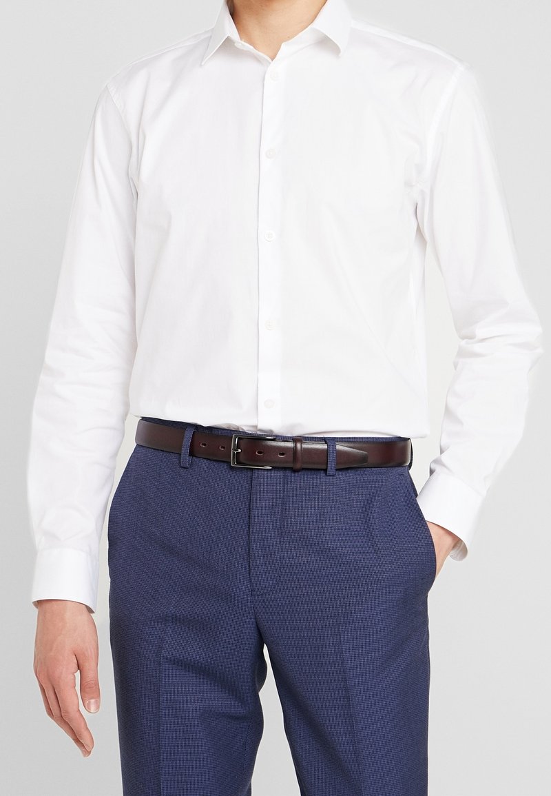 Homme portant une chemise blanche à manches longues boutonnée, rentrée dans un pantalon bleu tailleur avec une ceinture en cuir marron foncé.