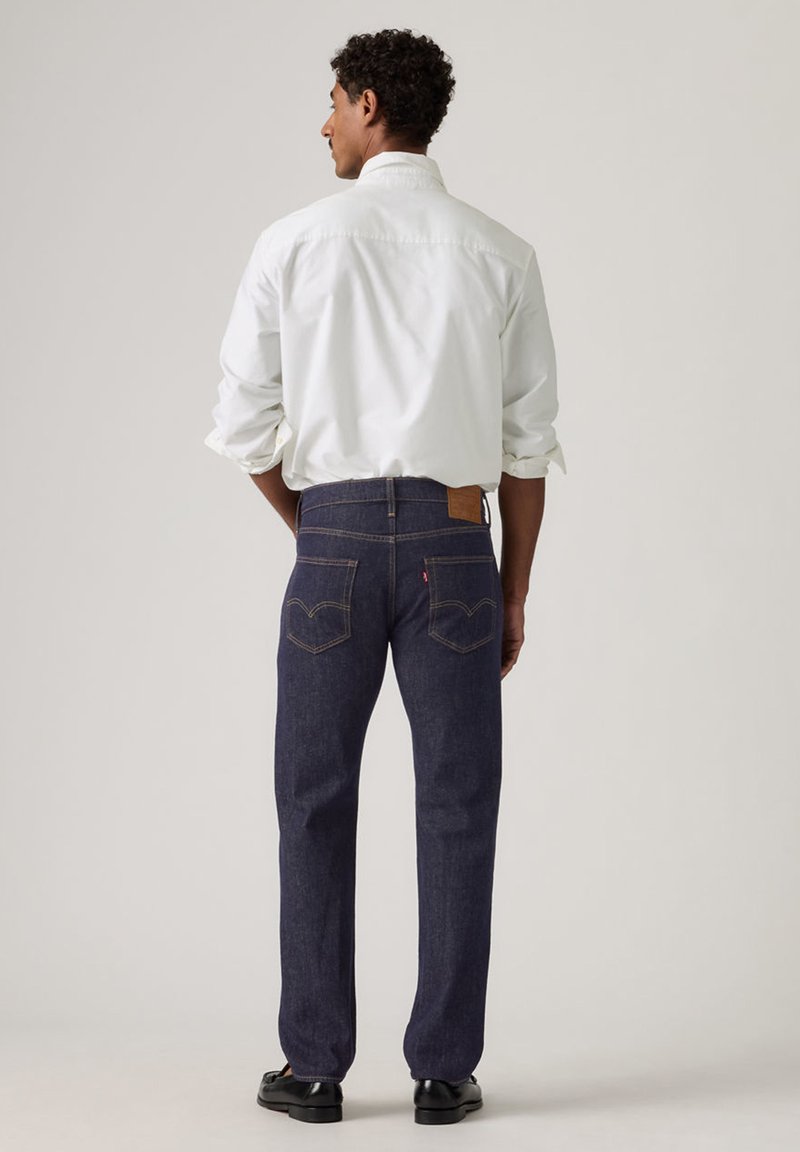 Hombre con vaqueros azul oscuro y una camisa blanca de manga larga, de pie con las manos en los bolsillos, de espaldas contra un fondo liso.